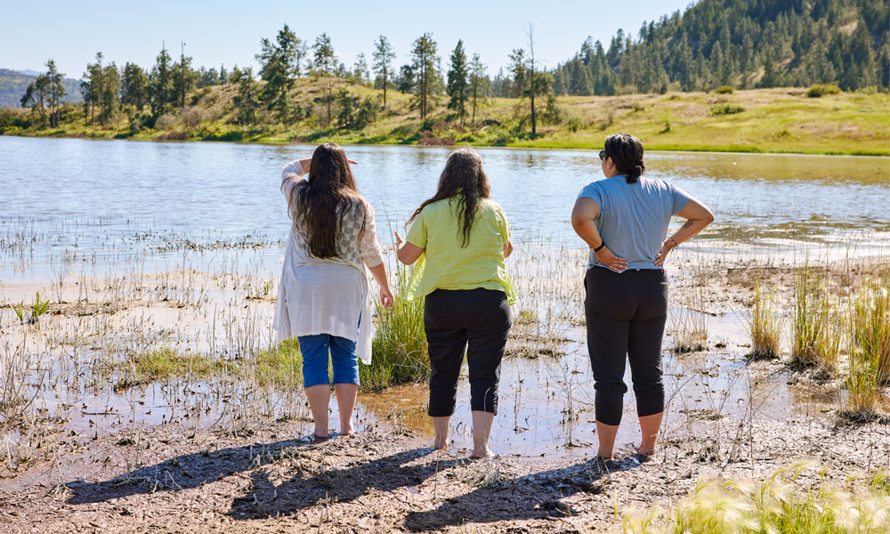 Three women stand looking at a lake, with their backs to the camera.